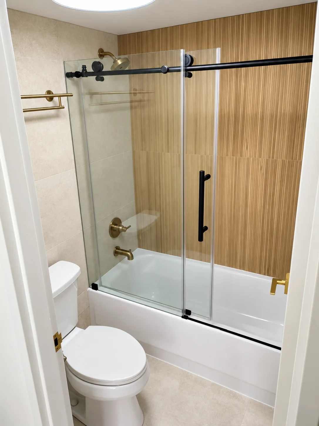 Warm-toned bathroom with fluted wood-look tile accent wall, sliding glass tub enclosure and brushed brass fixtures
